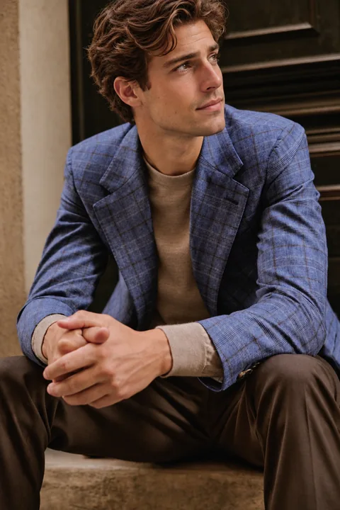 Sitzender Herr im blau karierten Sakko über braunem Strickpullover, Hände gefaltet, nachdenklicher Blick zur Seite