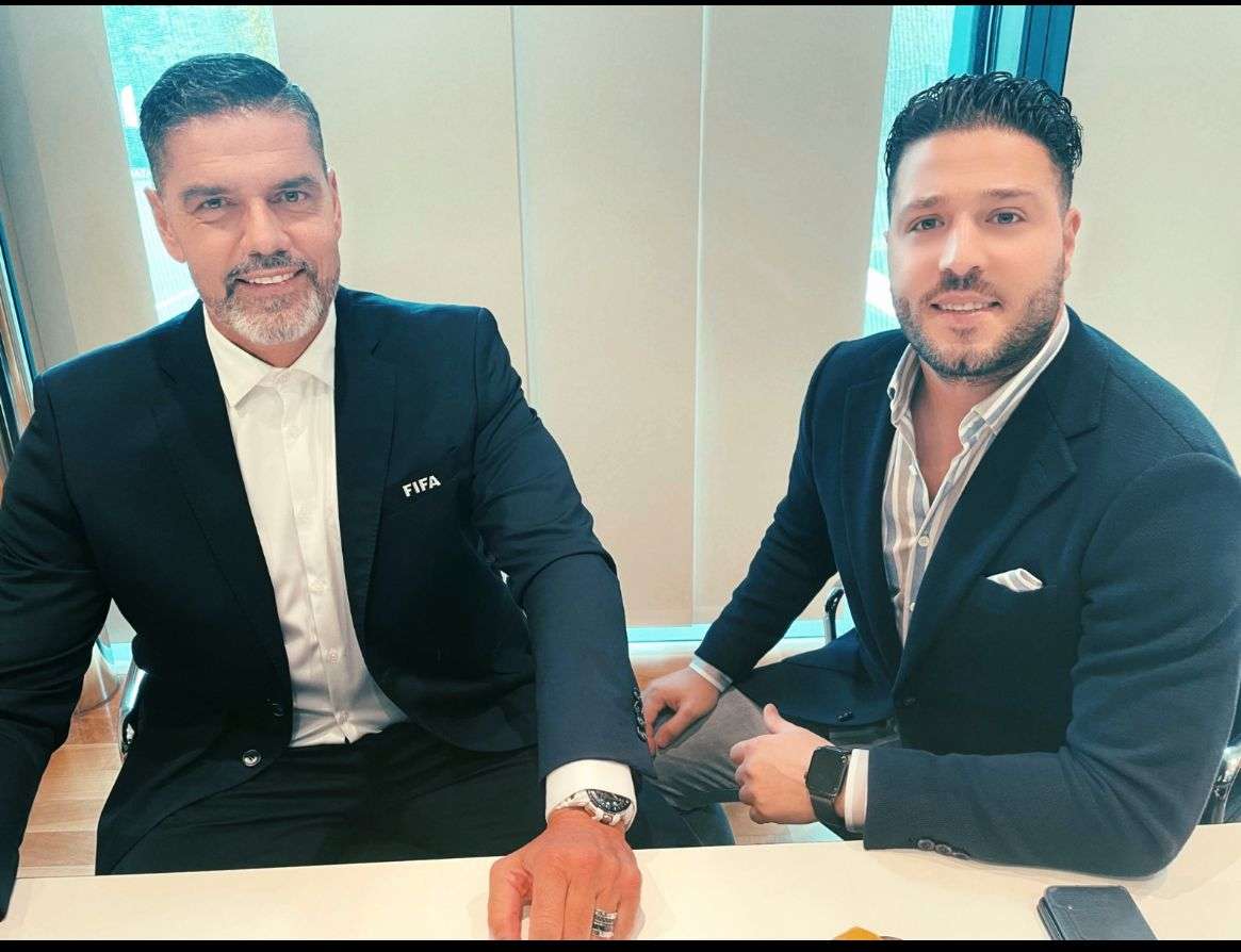 Zwei Männer in dunkelblauen Jacketts am Tisch, einer mit FIFA-Abzeichen am Revers