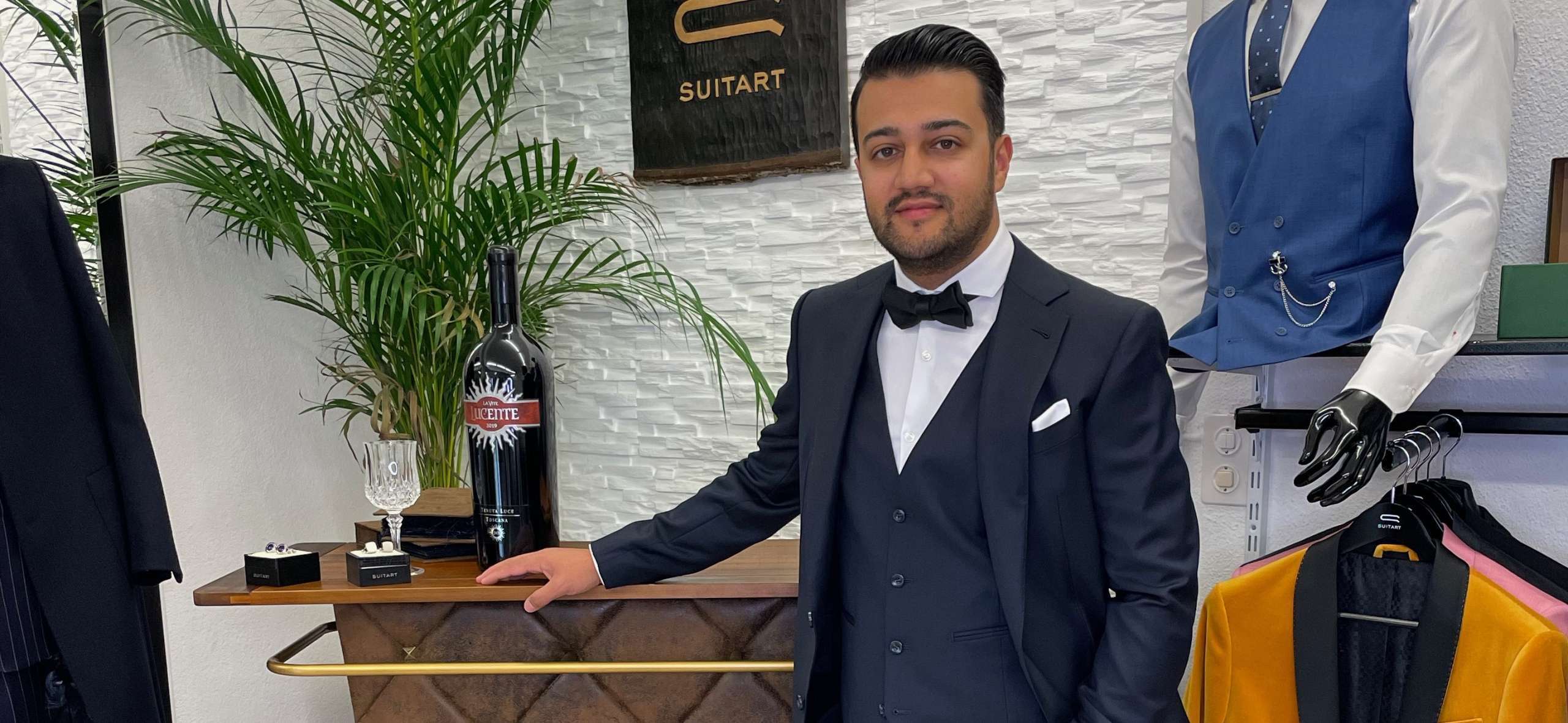 Mann im dunkelblauen Dreiteiler mit Fliege und Einstecktuch lehnt an der Bar im SuitArt Geschäft
