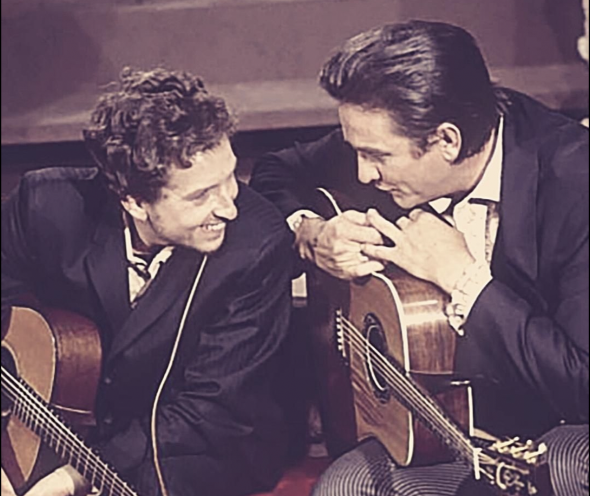 Johnny Cash und ein weiterer Musiker sitzen mit Gitarren nebeneinander und lachen, beide in dunklen Anzügen, in Sepia
