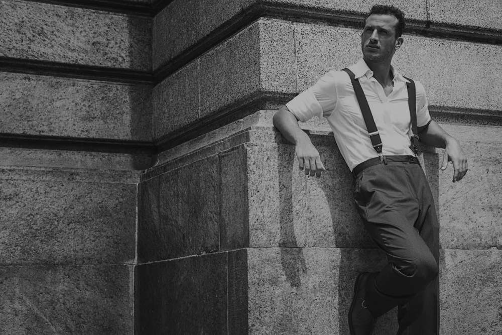 Uomo in camicia bianca, bretelle e pantaloni a vita alta appoggiato a un muro di pietra, in bianco e nero
